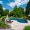 A luxurious backyard with a curvy swimming pool surrounded by lush greenery, Philly Travertine Pavers and Copings, lounge chairs, and umbrellas under a clear blue sky.