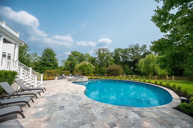 A luxurious backyard featuring a large, kidney-shaped swimming pool surrounded by a spacious stone patio with loungers, crafted from silver travertine pavers and copings, adjacent to a lush green lawn.