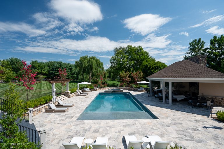 A luxurious backyard featuring a large rectangular swimming pool surrounded by silver travertine pavers and copings, sun loungers, a covered pavilion with comfortable seating, and lush greenery under a blue
