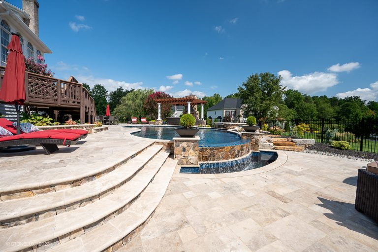A luxurious backyard featuring a curved pool with walnut travertine pavers and copings, a wooden deck with red lounge chairs, and lush landscaping under a clear blue sky.