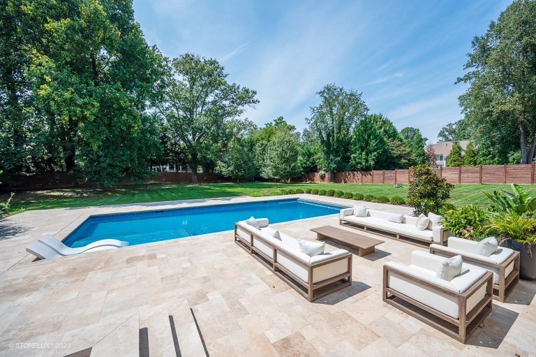 Backyard with a rectangular swimming pool surrounded by walnut travertine pavers and copings, white lounge furniture, a lush green lawn, and tall trees behind a wooden fence under a clear blue sky.