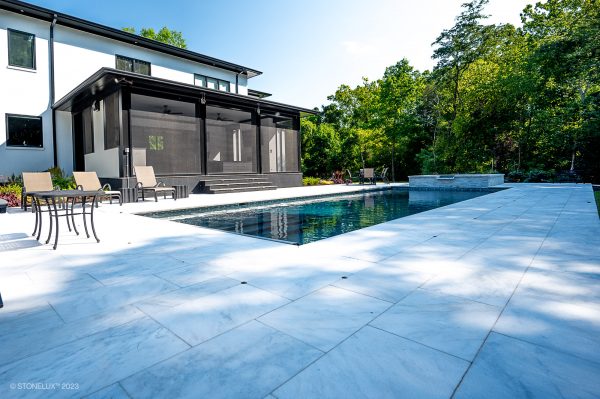 Luxurious backyard with a large swimming pool, surrounded by a spacious Afyon White Marble Sand Blasted French Pattern 3cm Paver tiled patio, modern two-story house, patio furniture, and lush trees under a clear blue sky.