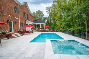 A luxurious backyard with a large swimming pool and a smaller attached hot tub, surrounded by Afyon White Marble Sand Blasted French Pattern 3cm Paver stone patio, complemented by red umbrellas and matching cushions on outdoor furniture.