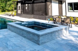 A modern outdoor setup with a small square hot tub on Afyon White Marble Sand Blasted copings, surrounded by patio furniture, near a pool and a contemporary house with large windows.