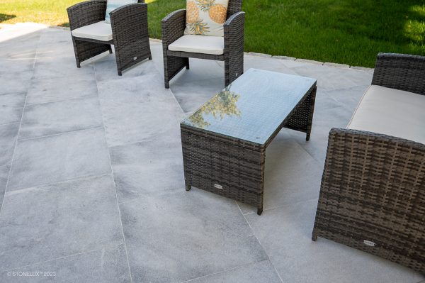 Outdoor patio setting with wicker armchairs, cushions, and a matching coffee table on Arctic Grey Marble Sand Blasted 24x24 3cm paver stone tiles, surrounded by a lush green lawn.