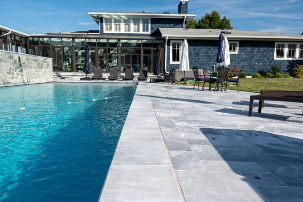 Luxurious backyard with a large swimming pool, Arctic Grey Marble Sand Blasted copings, and modern poolside furniture, set against a sprawling house with extensive glass paneling under a clear blue sky.