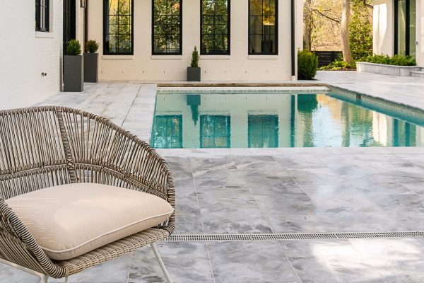 A luxurious outdoor pool beside a modern house, featuring a large wicker lounge chair on an Arctic Grey Marble Sand Blasted Paver deck, clear blue water, and lush greenery in the background.