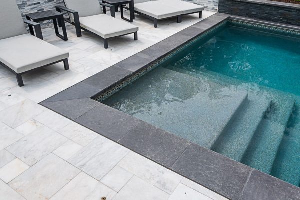 Modern outdoor pool area with a blue swimming pool surrounded by gray stone tiles, featuring Black Basalt Brushed Eased Edge Pool copings and adjacent lounge chairs and benches.