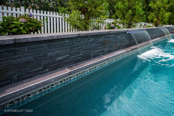 A serene outdoor swimming pool with clear blue water, featuring a Black Basalt Brushed Eased Edge Tread wall with decorative accents and multiple water jets creating arcs that fall into the pool. Lush greenery surrounds the area.