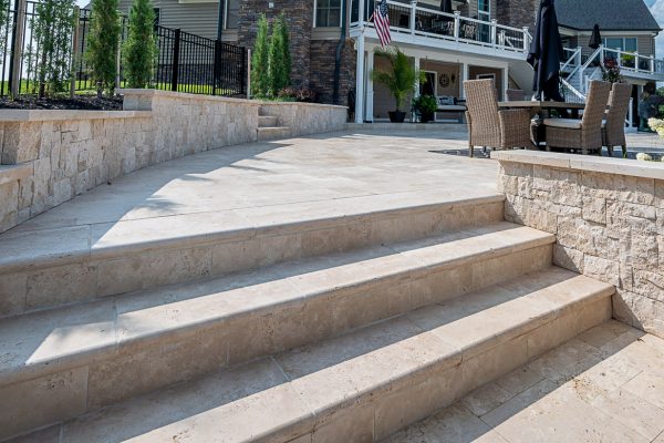 An outdoor patio area featuring large stone steps with Ivory Travertine Tumbled 16"x24"x2" Double Bullnose Coping leading up to a deck with Ivory Travertine paving, connected to a stately home with a visible American flag.