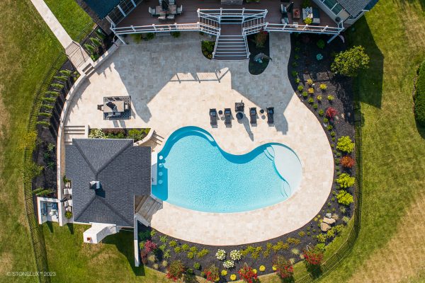Aerial view of a backyard featuring a kidney-shaped swimming pool surrounded by a spacious patio with Ivory Travertine Tumbled 24x24 3cm pavers and well-manicured gardens adjacent to a residential home.