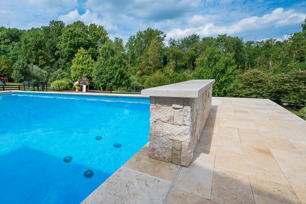 A bright, sunny day at an outdoor pool with clear blue water. A modern diving platform made of Ivory Travertine Tumbled 12"x24"x2" Single Bullnose Coping - Premium extends over the pool, surrounded by lush green trees.