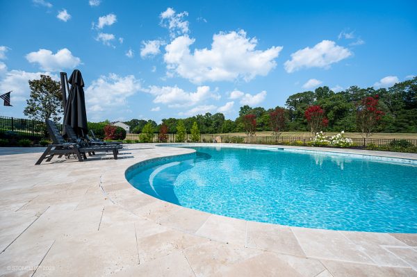 A curvy outdoor swimming pool with pristine, blue water, surrounded by Ivory Travertine Tumbled French Pattern 3cm Paver deck with black lounge chairs, overlooking a scenic view of trees and a clear sky.