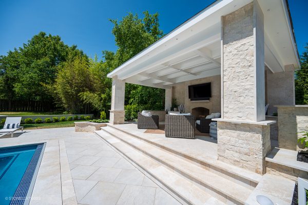 An elegant outdoor patio area beside a pool, featuring a covered seating area with a fireplace, modern wicker furniture, and Pera Cream Marble Sand Blasted Eased Edge Treads, set against a backdrop of lush green trees.