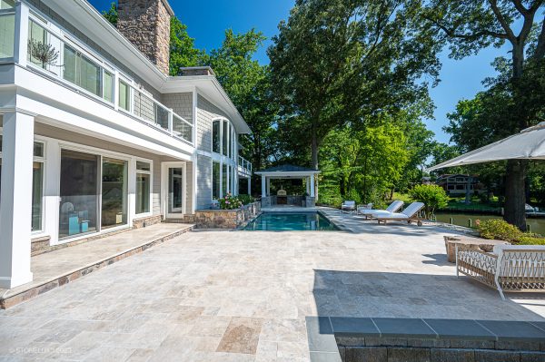 Luxurious backyard with a swimming pool, sun loungers, and a spacious patio featuring Philly Travertine Tumbled Paver. It also includes a large, well-maintained.
