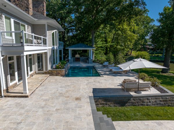 A luxurious backyard featuring a stone patio with a Philly Travertine Tumbled swimming pool deck, surrounded by lush greenery, a pavilion, and upscale outdoor furniture. The house is large with white exteriors and multiple balconies.