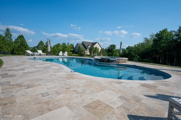 An expansive outdoor swimming pool with clear, calm water, surrounded by a large stone patio made from Philly Travertine Tumbled French Pattern 3cm Pavers and featuring a nearby waterfall and several statues, under a clear blue.