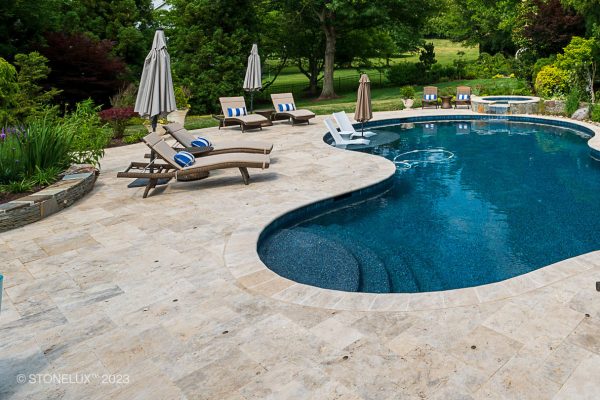 An elegant backyard pool area with curved edges, surrounded by Philly Travertine Tumbled Pavers. Poolside loungers with white umbrellas and lush greenery enhance the serene atmosphere.