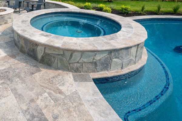 A round, stone-walled jacuzzi featuring Silver Travertine Tumbled 6"x12"x3CM Single Bullnose Coping, connected to a larger swimming pool with blue water, and surrounded by a tiled patio in a landscaped backyard.
