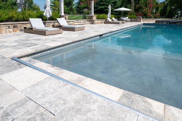 A serene outdoor pool with clear blue water, surrounded by a Silver Travertine Tumbled 12x24x2 Single Bullnose Coping - Premium deck and two lounge chairs under white umbrellas, set in a lush green landscape.