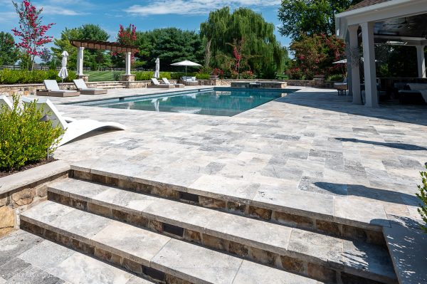 A luxurious backyard with a rectangular swimming pool surrounded by Silver Travertine Tumbled 24x72x2 Eased Edge Tread - Premium coping, featuring green landscaped gardens and outdoor structures under a clear blue sky.