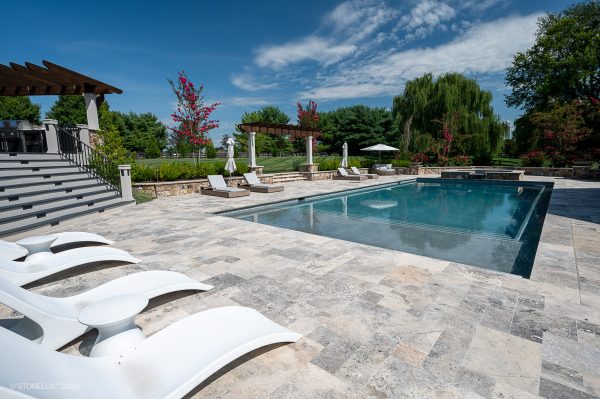 An elegant outdoor swimming pool with clear blue water, surrounded by a silver travertine patio and accompanied by a covered seating area and sun loungers, set amidst lush green landscaping under a clear sky.