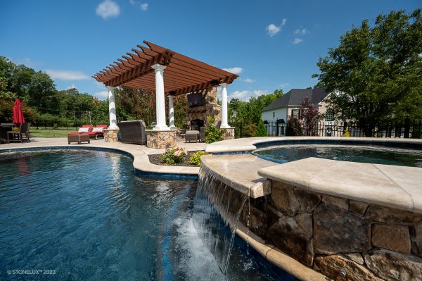 A luxurious backyard with a curved pool featuring a waterfall and stone edges, a pergola with seating and a TV, surrounded by lush greenery under a clear blue sky. The pool boasts elegant Walnut Travertine Tumbled 24
