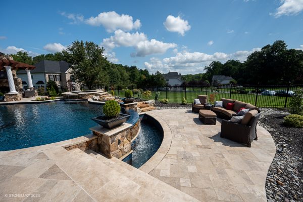 Luxurious backyard with a swimming pool featuring waterfalls and integrated fire pits, surrounded by a Walnut Travertine Tumbled 24"x72"x2" Eased Edge Tread - Premium patio and plush seating, lush greenery, and a clear sky above.