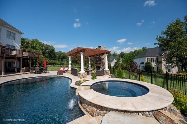 A luxurious backyard with a curved swimming pool, a hot tub, and a covered patio area with lounge chairs under a pergola. Lush green lawn and neighboring houses visible in the background under a clear Walnut Travertine Tumbled 24