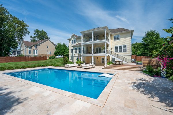 A luxurious backyard featuring a large swimming pool with clear blue water, surrounded by a sandstone patio upgraded with Walnut Travertine Tumbled 16