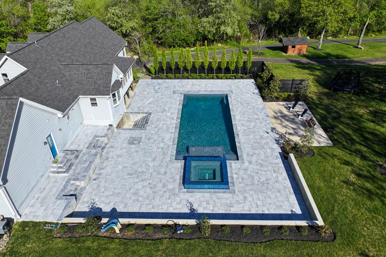 Aerial view of a backyard with a large rectangular blue swimming pool, surrounded by an auto-draft patio, adjacent to two homes and a green lawn area with trees.