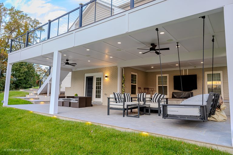 A spacious covered porcelain paver patio in Fairfax features modern outdoor furniture, including a hanging bench, two ceiling fans, recessed lighting, and a wall-mounted TV. The area is framed by white pillars and overlooks a lawn. A staircase leads to an upper-level deck with railing.