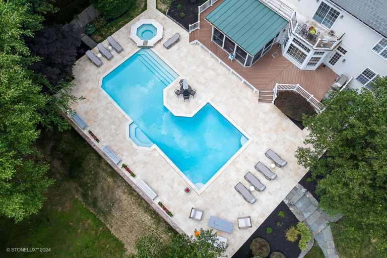 Aerial view of a luxurious backyard featuring a large hexagonal swimming pool bordered by an Ivory Travertine pool deck, multiple lounge chairs, a shaded seating area with umbrellas, and a separate circular hot tub attached. The pool area is surrounded by lush greenery and a modern white house.