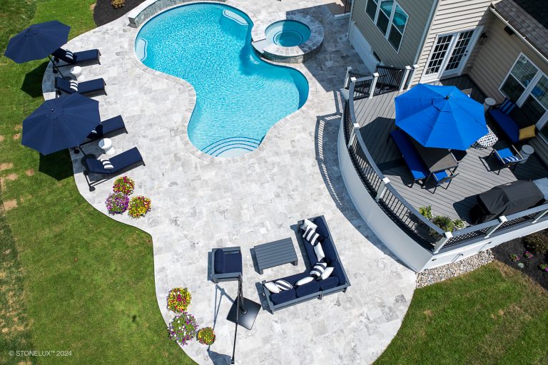 A spacious backyard with a kidney-shaped swimming pool, an adjacent hot tub, and a patio featuring lounge chairs, outdoor sofas, and blue umbrellas set on Tundra Grey Marble Sandblasted tiles. Brightly colored potted flowers add vibrant accents to the stone-tiled patio. A deck overlooks the pool area.