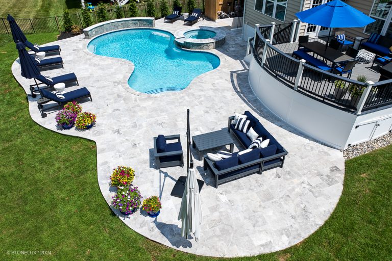 A spacious backyard with a kidney-shaped swimming pool, an adjacent hot tub, and a patio featuring lounge chairs, outdoor sofas, and blue umbrellas set on Tundra Grey Marble Sandblasted tiles. Brightly colored potted flowers add vibrant accents to the stone-tiled patio. A deck overlooks the pool area.