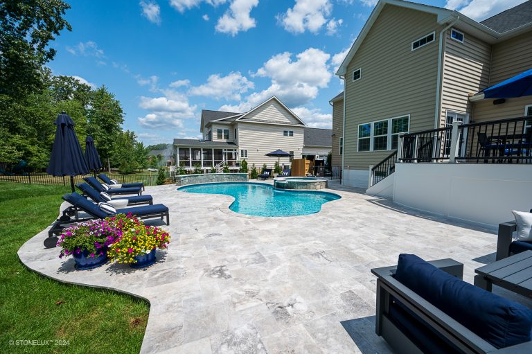 A spacious backyard with a kidney-shaped swimming pool, an adjacent hot tub, and a patio featuring lounge chairs, outdoor sofas, and blue umbrellas set on Tundra Grey Marble Sandblasted tiles. Brightly colored potted flowers add vibrant accents to the stone-tiled patio. A deck overlooks the pool area.