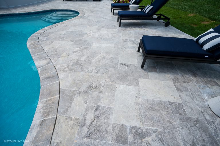 A spacious backyard with a kidney-shaped swimming pool, an adjacent hot tub, and a patio featuring lounge chairs, outdoor sofas, and blue umbrellas set on Tundra Grey Marble Sandblasted tiles. Brightly colored potted flowers add vibrant accents to the stone-tiled patio. A deck overlooks the pool area.