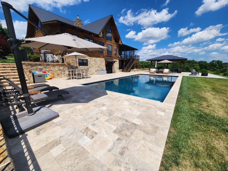 A luxurious outdoor scene features a stone patio of Philly Travertine surrounding a rectangular pool. Two umbrellas offer shade over seating areas. A wooden house with large windows is in the background, set against a bright blue sky with scattered clouds.